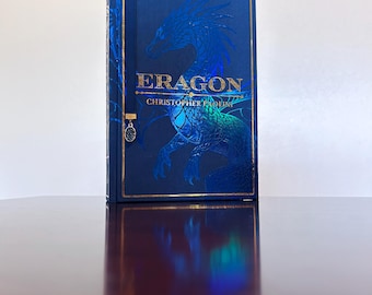 May include: A hardcover book titled Eragon by Christopher Paolini with a deep blue cover featuring a large, embossed dragon illustration in metallic blue and gold foil accents, complete with a dark blue ribbon bookmark and a small oval charm.