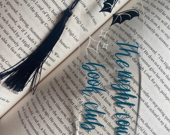 May include: A clear acrylic bookmark with a teal and white design of an open book with wings. The bookmark features the text "The High Lords" in a teal script font.