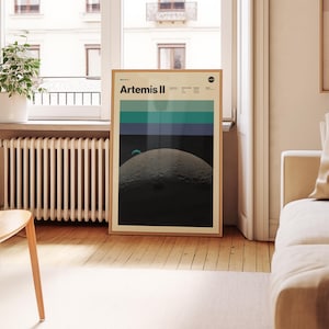 May include: A framed poster featuring the text "Artemis II" at the top. The poster has a design of horizontal stripes in shades of teal, blue, and black above a depiction of a moon's surface. The poster is in a light brown wooden frame.