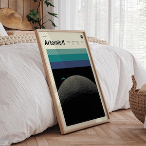 May include: A framed poster with the text "Artemis II" features a graphic of a planet and a color block design in shades of blue and green. The poster is in a light wood frame and leans against a white bedspread.