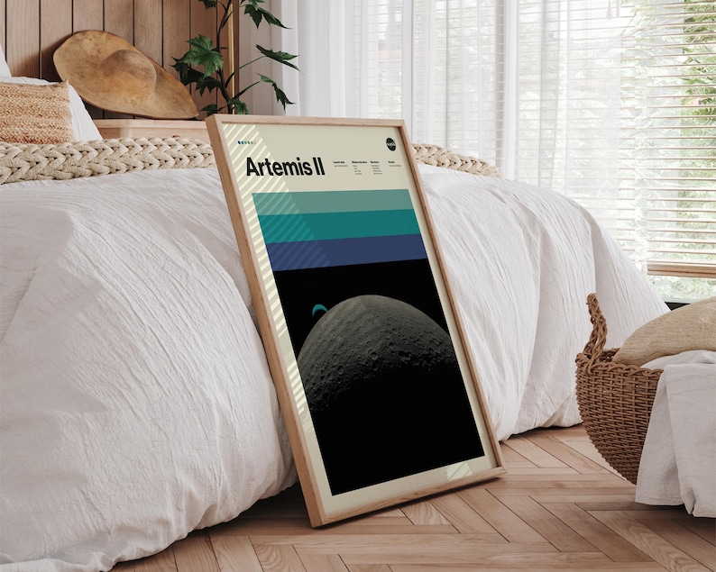 May include: A framed poster with the text "Artemis II" features a graphic of a planet and a color block design in shades of blue and green. The poster is in a light wood frame and leans against a white bedspread.