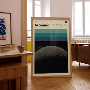 May include: A framed poster with the text "Artemis II" and a NASA logo. The poster features a graphic of the moon and a color block design in shades of blue and green. The poster is in a light wood frame.