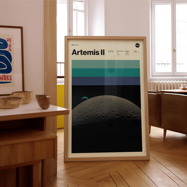 May include: A framed poster with the text "Artemis II" and a NASA logo. The poster features a graphic of the moon and a color block design in shades of blue and green. The poster is in a light wood frame.