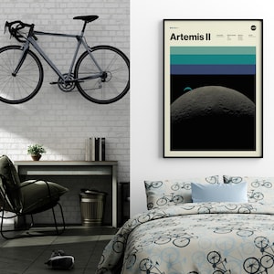 May include: A gray road bicycle mounted on a white brick wall. The bicycle has black handlebars, wheels, and a black seat. A framed poster with the text "Artemis II" hangs on the wall. A desk, chair, and bed with bicycle-themed bedding are also visible.