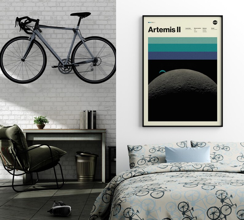 May include: A gray road bicycle mounted on a white brick wall. The bicycle has black handlebars, wheels, and a black seat. A framed poster with the text "Artemis II" hangs on the wall. A desk, chair, and bed with bicycle-themed bedding are also visible.