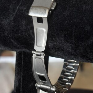 May include: A silver metal watch band with a clasp and links. The band is on a black velvet display stand.
