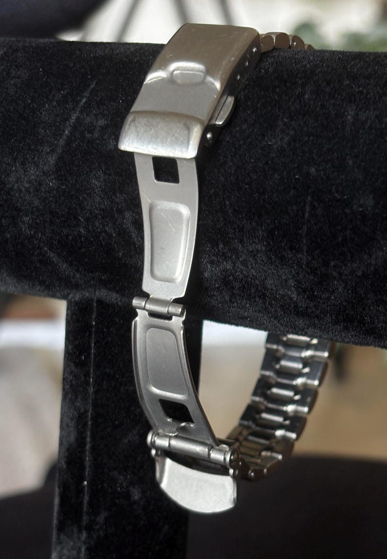 May include: A silver metal watch band with a clasp and links. The band is on a black velvet display stand.