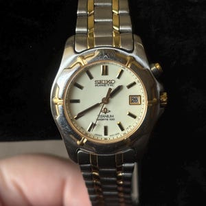 May include: A silver and gold two-tone Seiko Kinetic watch with a white face and black hands. The watch has a date window at the 3 o'clock position and the words "Titanium Sports 100" printed on the face.