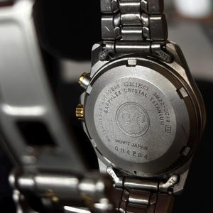 May include: Back of a silver Seiko watch with a black face. The watch is made of titanium and has a sapphire crystal. The watch is water resistant to 10 bar and has a movement made in Japan. The watch has the model number 3M22-0C49 and the serial number 6N4204.
