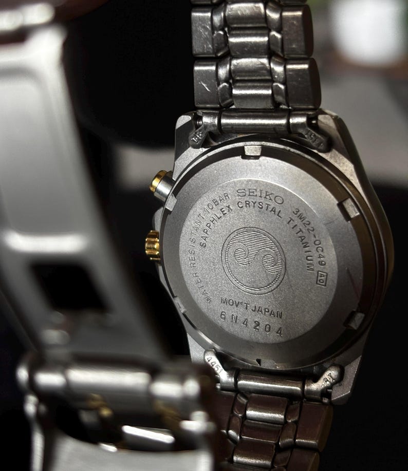 May include: Back of a silver Seiko watch with a black face. The watch is made of titanium and has a sapphire crystal. The watch is water resistant to 10 bar and has a movement made in Japan. The watch has the model number 3M22-0C49 and the serial number 6N4204.