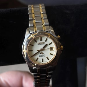 May include: A two-tone gold and silver Seiko Kinetic watch with a white face and a date window. The watch has a titanium band and says "Titanium Sports 100" on the face.