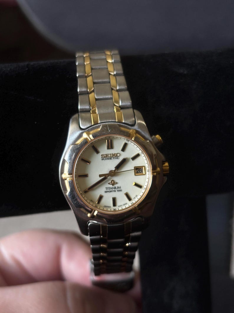 May include: A two-tone gold and silver Seiko Kinetic watch with a white face and a date window. The watch has a titanium band and says "Titanium Sports 100" on the face.