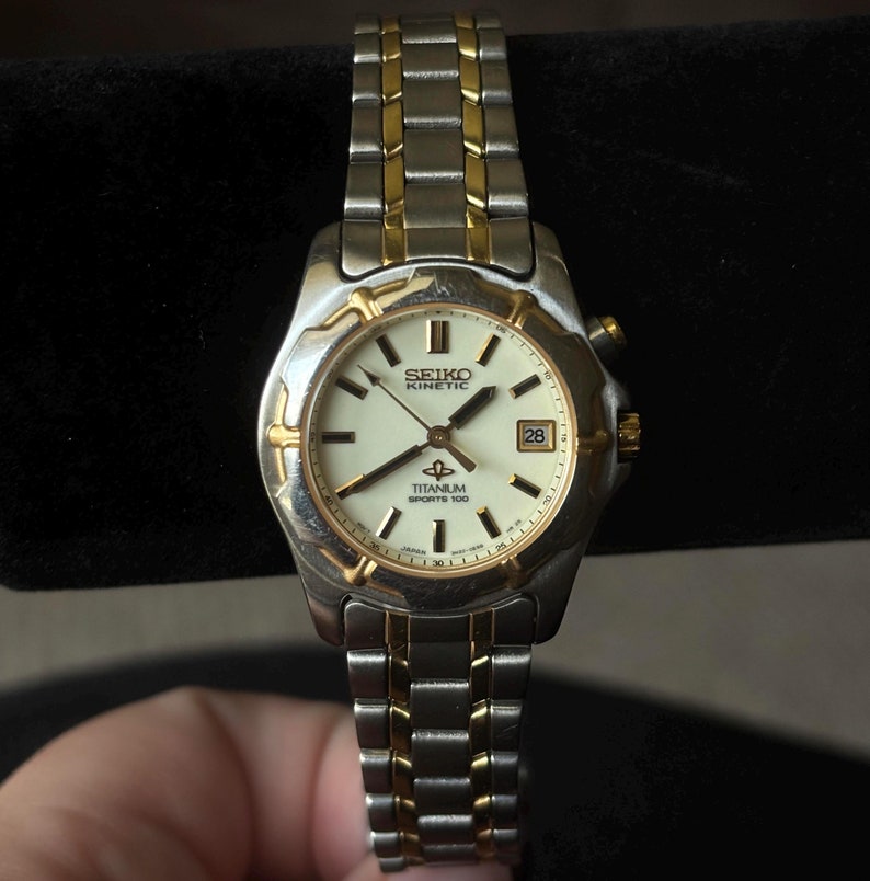 May include: A two-tone silver and gold Seiko Kinetic watch with a white face. The watch face has black markings and the text "SEIKO KINETIC" and "TITANIUM SPORTS 100". The date window shows "28".