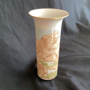 May include: A white ceramic vase with a gold rim and a painted scene of a desert landscape with brown rocks and green plants.