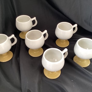 May include: Six white ceramic cups with handles, each set on a small, round, tan base. The cups have a rounded shape and are arranged on a dark background. The cups are likely for serving beverages.