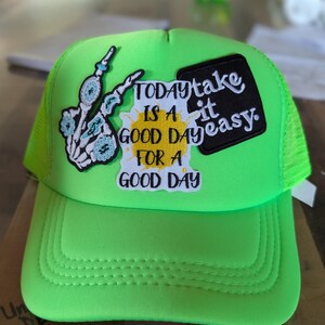 Pode incluir: Um boné trucker verde neon com três emblemas bordados. Um emblema mostra uma mão esquelética com detalhes em turquesa, outro exibe o texto amarelo Today is a good day for a good day, e um emblema preto traz a inscrição take it easy.