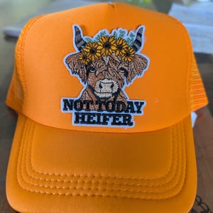 Pode incluir: Um boné trucker laranja brilhante com um emblema bordado de uma vaca Highland castanha a usar uma coroa de girassóis. O emblema inclui o texto preto "NOT TODAY HEIFER" posicionado diretamente abaixo da cara da vaca no painel frontal.