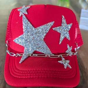 Pode incluir: Um boné trucker vermelho brilhante com parte traseira em rede e painel frontal sólido. A frente é decorada com várias aplicações de estrelas incrustadas com strass prateados de vários tamanhos e uma corrente metálica prateada na aba.