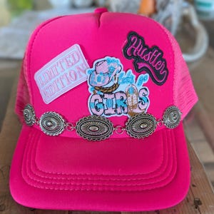Pode incluir: Um boné trucker rosa brilhante com uma corrente de conchos ovais prateados na frente. Os emblemas incluem uma etiqueta retangular rosa 'LIMITED EDITION', um emblema preto com a palavra 'Hustler' e um gráfico central com um chapéu de cowboy e 'Let's go GIRLS'.