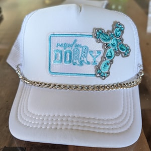 Pode incluir: Um boné trucker branco com um emblema retangular contendo o texto bordado em azul claro "raised on DOLLY" e uma cruz turquesa. Uma corrente de metal prateado está fixada na base do painel frontal, logo acima da aba do boné.