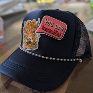 Pode incluir: Um boné trucker preto com parte traseira em rede e aba curva. O painel frontal apresenta um emblema bordado de uma vaca castanha adornada com girassóis amarelos e um emblema de balão de fala rosa com o texto You are beautiful. Um fio de pérolas brancas acentua a base do painel frontal.