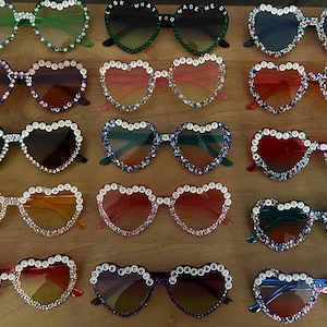 May include: A collection of 12 pairs of heart-shaped sunglasses with rhinestones and the words "Look What You Made Me Do" on the frames. The sunglasses are in various colors, including green, purple, red, orange, and blue.