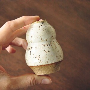 May include: A small, white ceramic vase with brown speckles. The vase has a rounded, bulbous shape and a slightly flared opening.