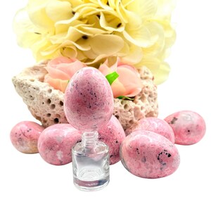 May include: A collection of pink, speckled, egg-shaped stones with a small, clear glass bottle. The stones are arranged with a backdrop of a porous, beige rock formation and soft yellow and pink flowers. The composition is set against a white background.