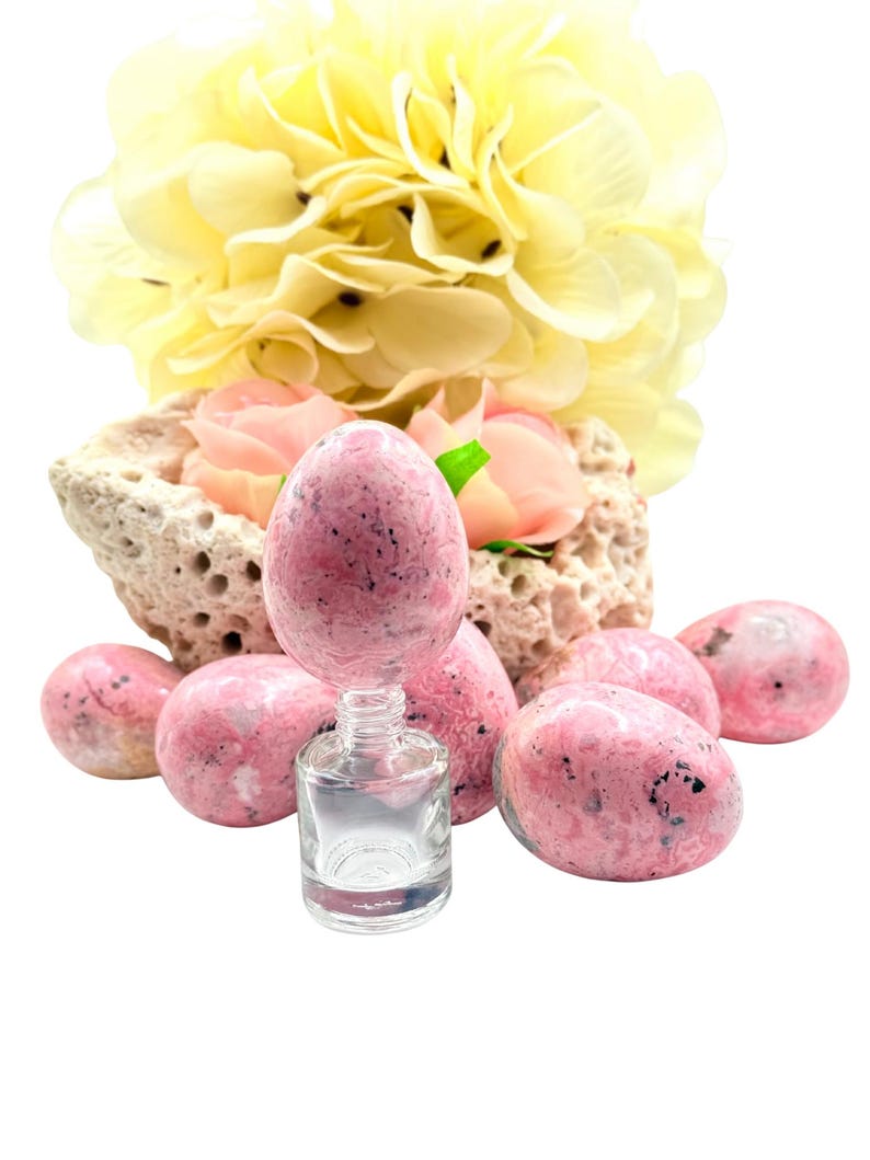 May include: A collection of pink, speckled, egg-shaped stones with a small, clear glass bottle. The stones are arranged with a backdrop of a porous, beige rock formation and soft yellow and pink flowers. The composition is set against a white background.