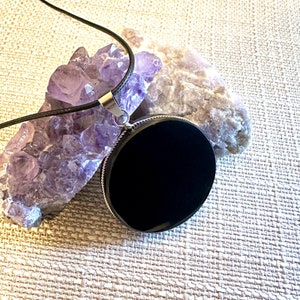May include: A black onyx pendant on a black cord necklace. The pendant is round and has a silver bezel. The necklace is resting on a bed of amethyst crystals.