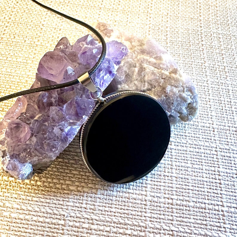May include: A black onyx pendant on a black cord necklace. The pendant is round and has a silver bezel. The necklace is resting on a bed of amethyst crystals.