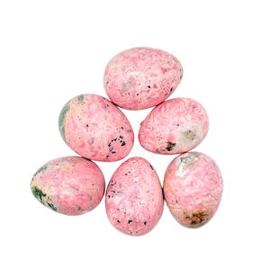 May include: A collection of six polished, egg-shaped stones. The stones are primarily pink with a marbled appearance, featuring darker pink, gray, and black speckles. Some stones have hints of green. The arrangement is a loose pyramid shape against a white background.