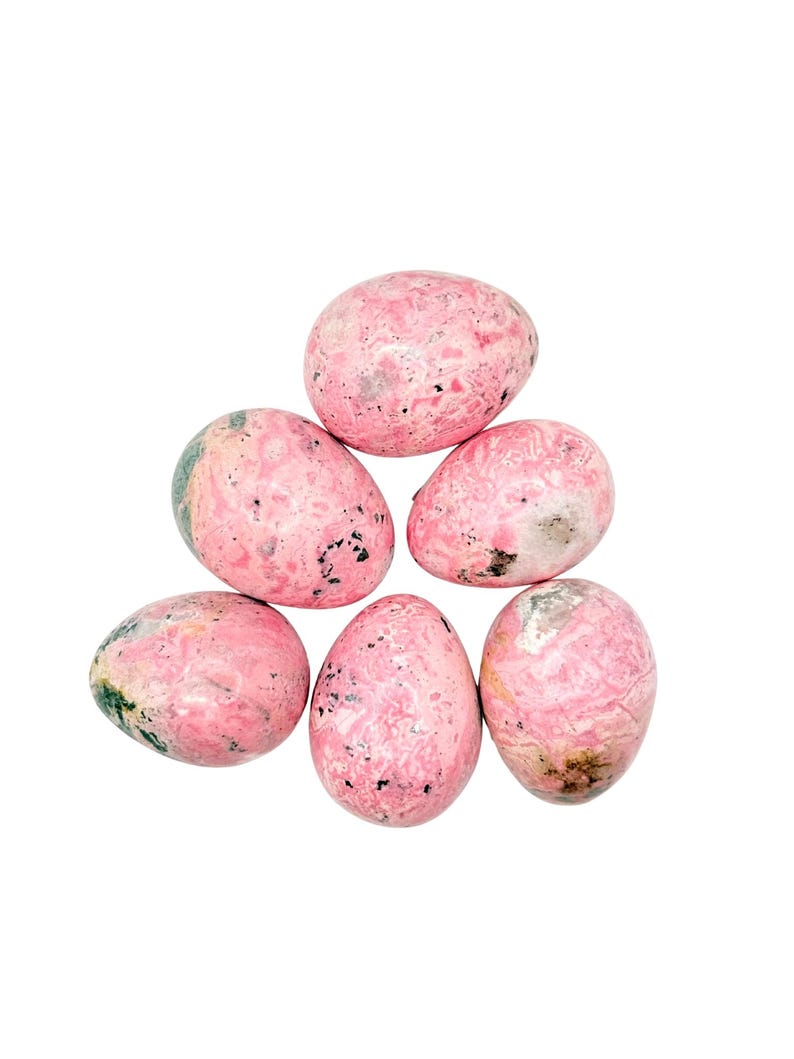 May include: A collection of six polished, egg-shaped stones. The stones are primarily pink with a marbled appearance, featuring darker pink, gray, and black speckles. Some stones have hints of green. The arrangement is a loose pyramid shape against a white background.
