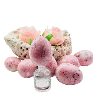May include: A collection of pink and black speckled stone eggs, some standing and some lying down. A small glass bottle with clear liquid is in the center, with a stone and pink roses in the background.