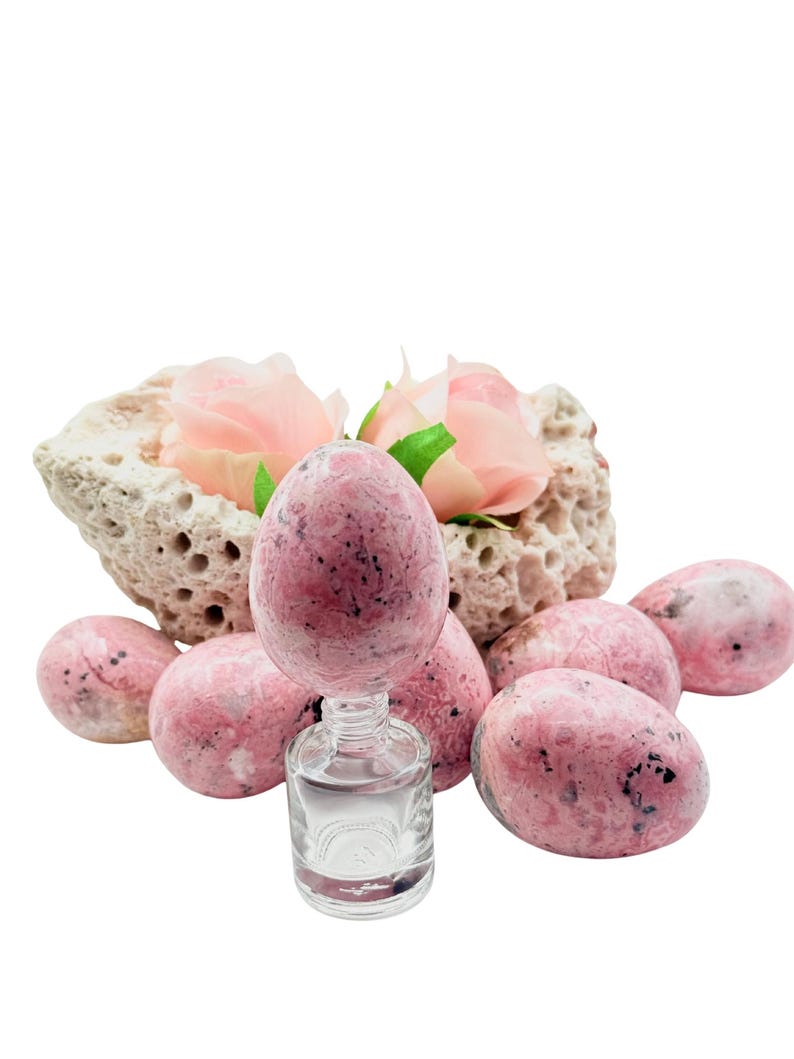 May include: A collection of pink and black speckled stone eggs, some standing and some lying down. A small glass bottle with clear liquid is in the center, with a stone and pink roses in the background.