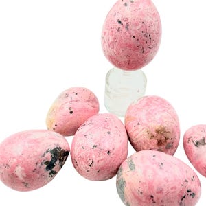 May include: Pink and black speckled egg-shaped stones. The stones are a light pink color with darker pink and black markings. The stones are smooth and polished, and appear to be made of a natural material. The stones are arranged in a group.
