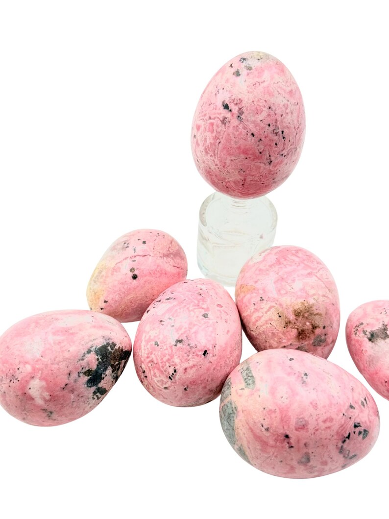 May include: Pink and black speckled egg-shaped stones. The stones are a light pink color with darker pink and black markings. The stones are smooth and polished, and appear to be made of a natural material. The stones are arranged in a group.