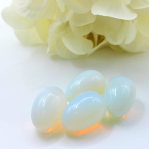 Puede incluir: Cuatro piedras ovaladas y opalescentes con un brillo blanco nacarado. Las piedras son lisas y pulidas, con un brillo sutil. Un grupo de pétalos de flores suaves de color crema se encuentra en el fondo.