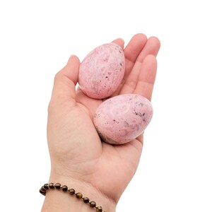 May include: Two pink, egg-shaped stones with speckled patterns are held in an open hand. The stones have a smooth, polished appearance. A beaded bracelet with brown beads and gold accents is visible on the wrist.