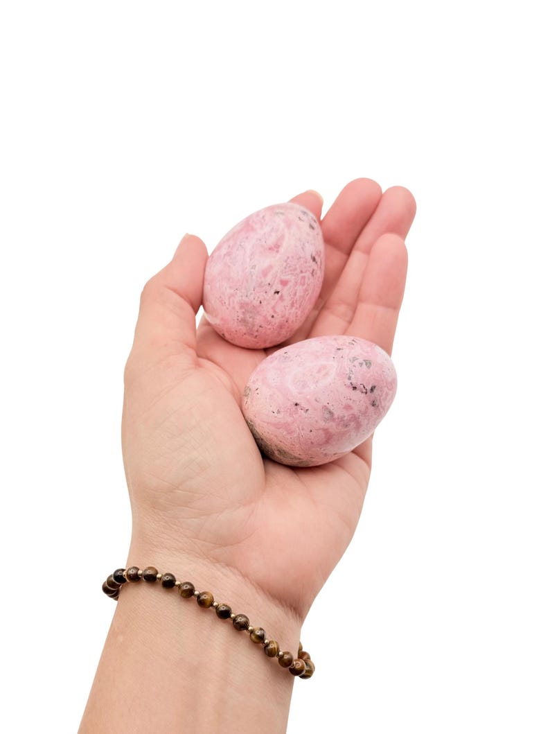 May include: Two pink, egg-shaped stones with speckled patterns are held in an open hand. The stones have a smooth, polished appearance. A beaded bracelet with brown beads and gold accents is visible on the wrist.