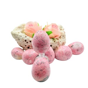May include: A collection of pink and gray stone eggs and spheres. One egg-shaped stone sits atop a small glass bottle. The stones are arranged around a porous, beige rock formation with pink roses.