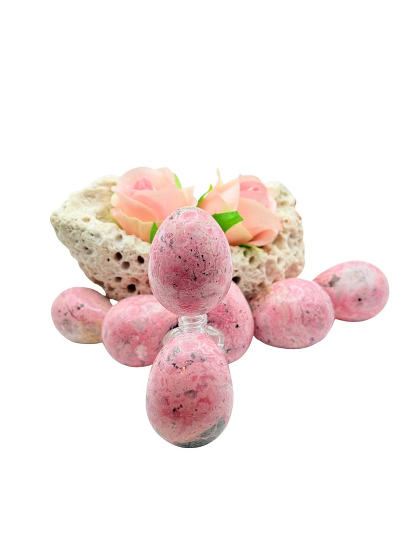 May include: A collection of pink and gray stone eggs and spheres. One egg-shaped stone sits atop a small glass bottle. The stones are arranged around a porous, beige rock formation with pink roses.