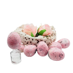 May include: A collection of pink and white speckled stone eggs, some resting near a small glass bottle and a porous, beige stone container holding two pink roses. The eggs vary in size and shape, creating a natural, organic aesthetic.