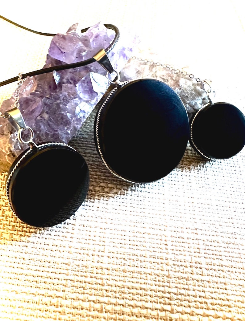 May include: Three black circular pendants of varying sizes on silver chains. The pendants are made of a smooth, polished material and have a sleek, modern look.