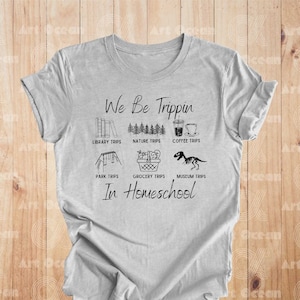 May include: A gray t-shirt with black text that reads "We Be Trippin' In Homeschool" and features a list of activities with icons, including library trips, nature trips, coffee trips, park trips, grocery trips, and museum trips.