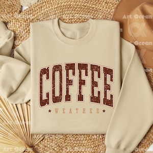 May include: A beige sweatshirt with the text "COFFEE WEATHER" in brown distressed lettering. The sweatshirt is laid out on a woven surface.
