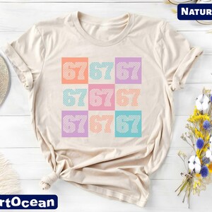 a beige t-shirt with a colorful graphic design of numbers on it, placed on a white surface alongside a hat and some flowers.