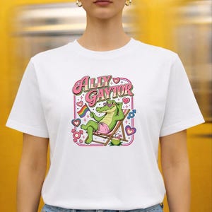 May include: A white t-shirt featuring a graphic print of a green alligator wearing pink shorts and purple sunglasses, lounging in a beach chair. The text reads "ALLY GAYTOR" above the alligator, surrounded by hearts, gender symbols, and a rainbow flag.