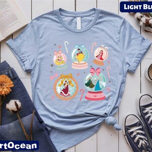 A light blue t-shirt with a colorful graphic of snowglows and various Disney princess characters, along with a pair of jeans, a book, a cup, and some flowers.