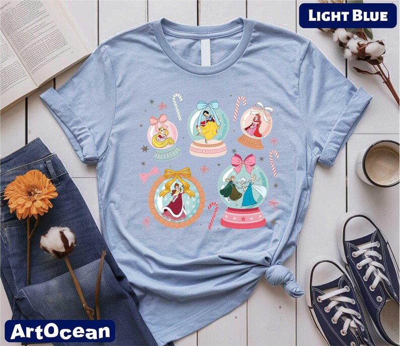 A light blue t-shirt with a colorful graphic of snowglows and various Disney princess characters, along with a pair of jeans, a book, a cup, and some flowers.